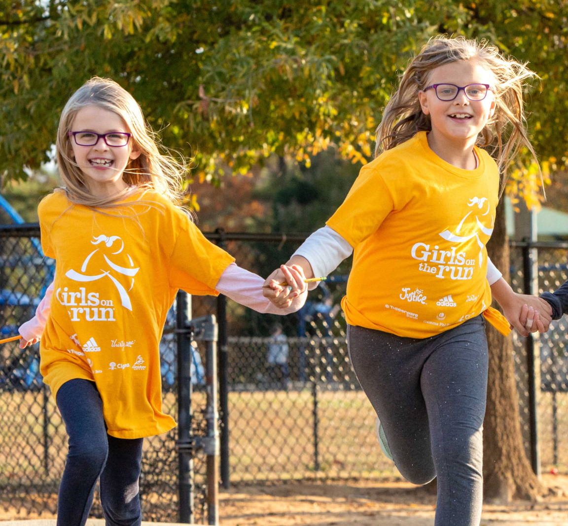 Two Girls on the Run participants in yellow shirts run while holding hands and smiling